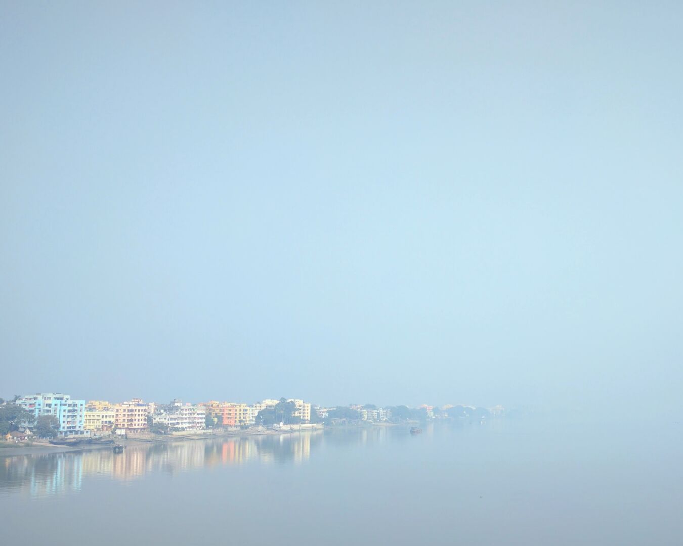 Photo of a shore of the river Ganges with buildings. The river reflects the sky and both meld into each other.